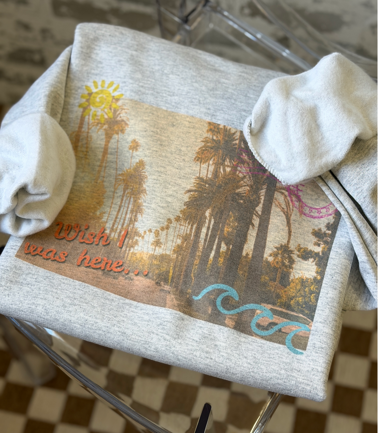 Gray sweatshirt featuring a vintage postcard-style palm tree scene with the phrase “wish I was here” in the bottom left corner