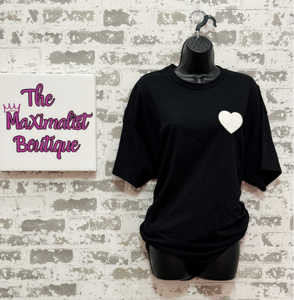 Black t-shirt with a white heart design on a mannequin against a textured wall with 'The Maximalist Boutique' branding.