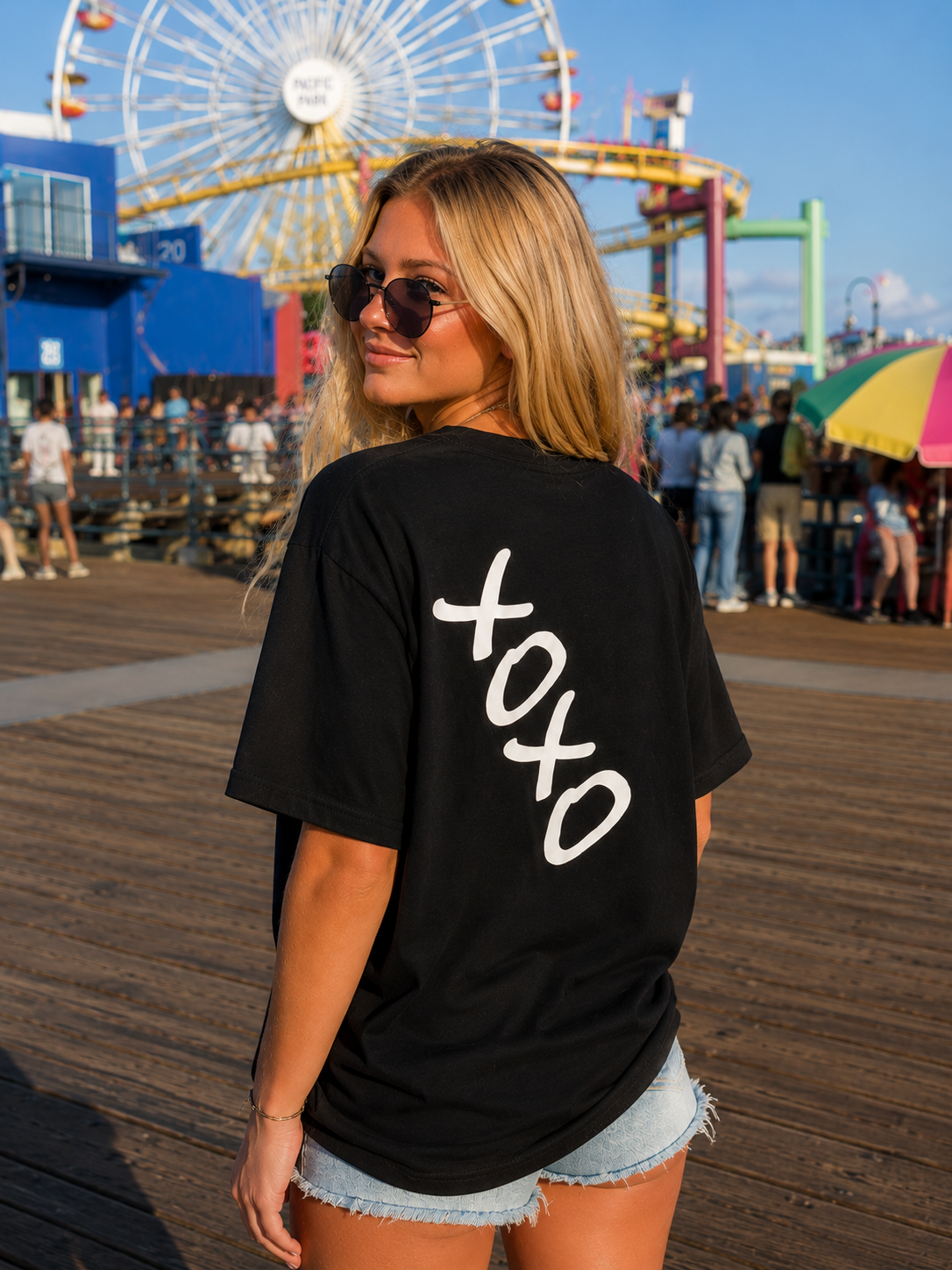 A woman on a wooden boardwalk wearing an oversized black t-shirt with 'xoxo' printed in white script down the back, with a Ferris wheel and pier crowd in the background.