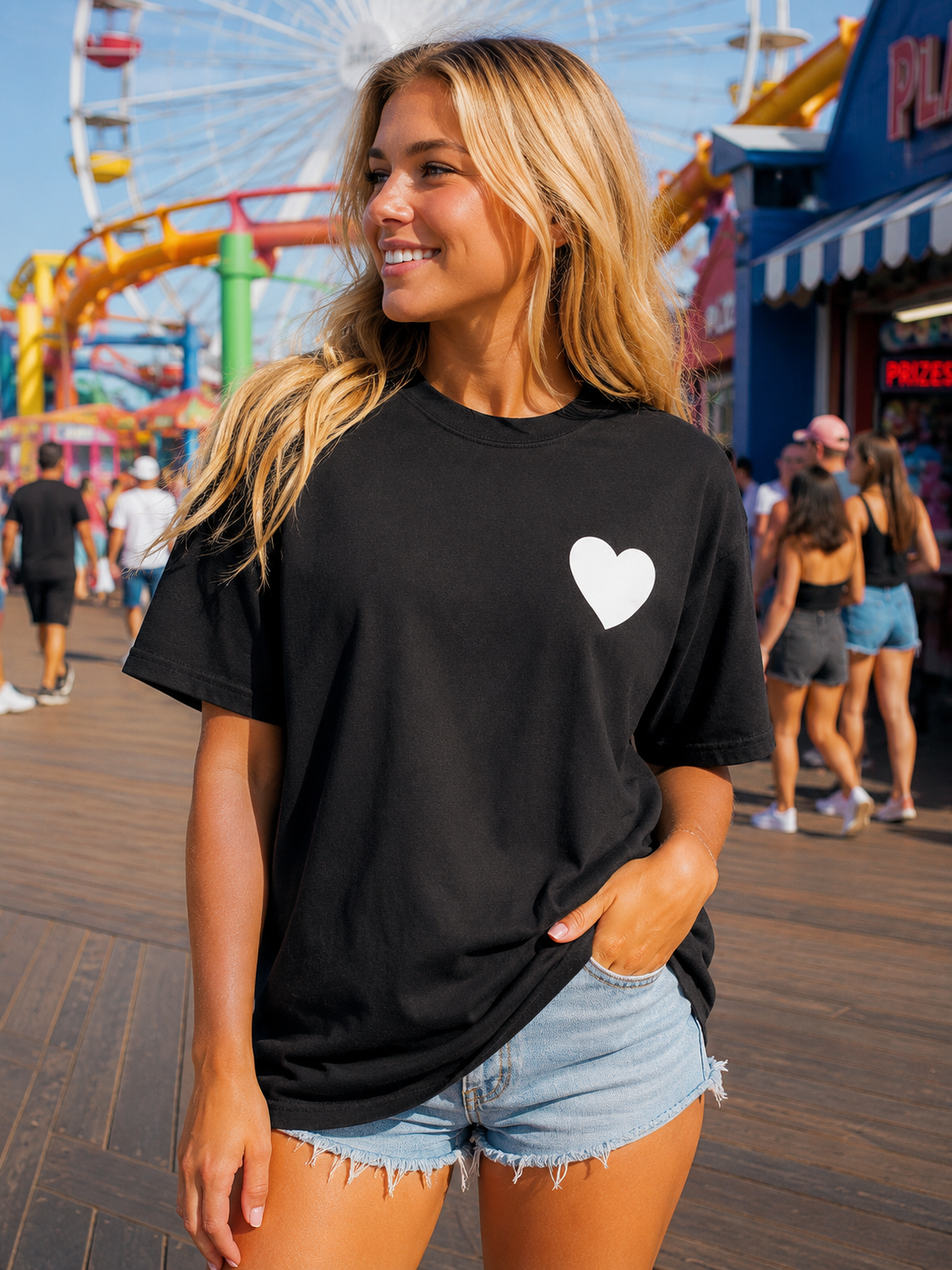 A woman wearing a black short-sleeve t-shirt with a small white heart on the left chest, standing on a wooden boardwalk in front of a colorful amusement park background.