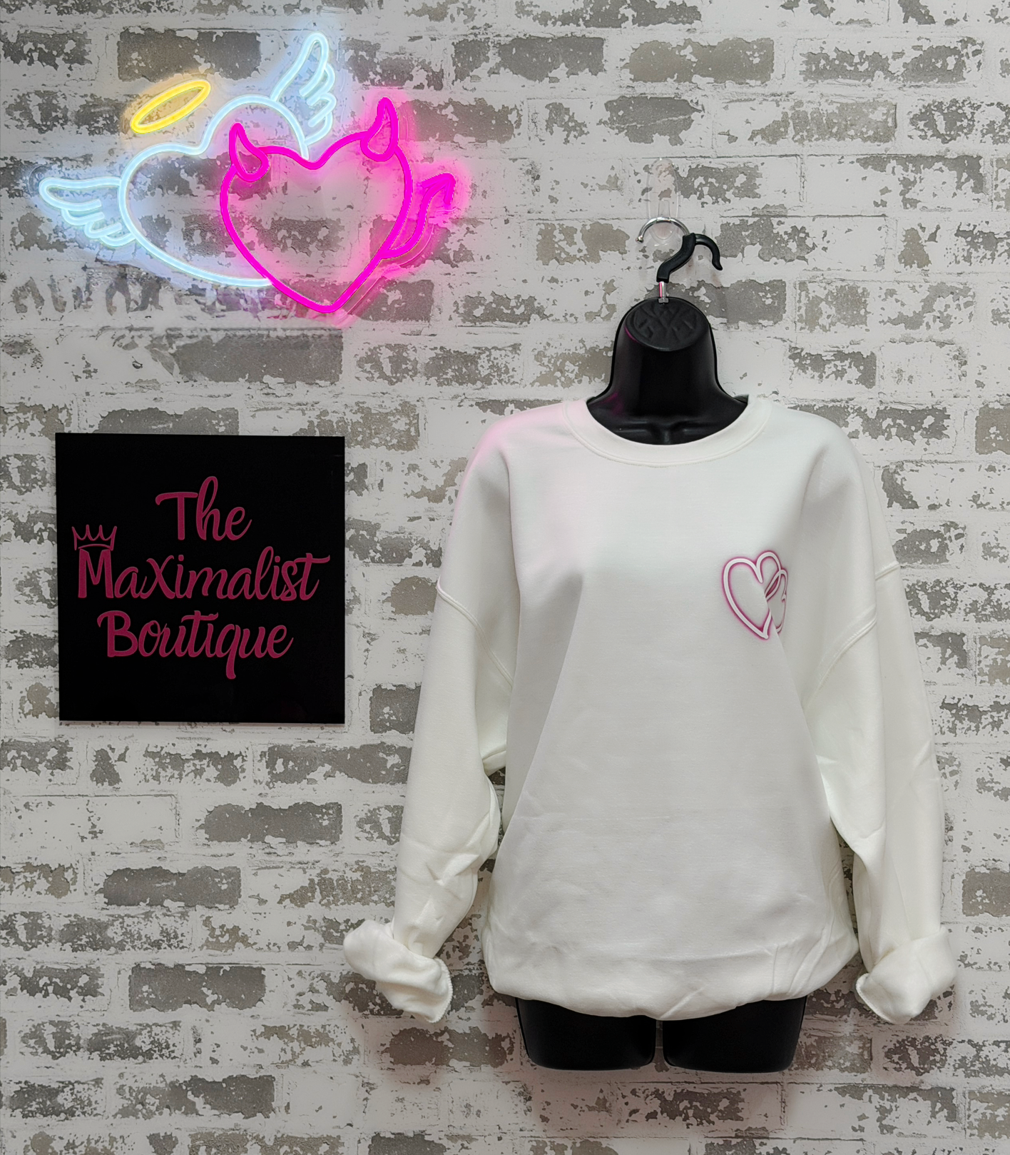 White sweatshirt with pink heart design on a mannequin against a textured wall with neon sign and 'The Maximalist Boutique' sign.