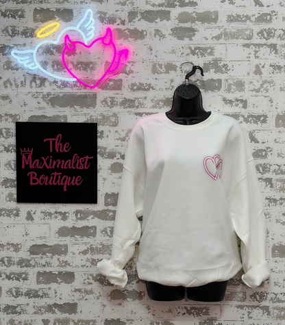 White sweatshirt with pink heart design on a mannequin against a textured wall with neon sign and 'The Maximalist Boutique' sign.