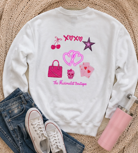 A white crewneck sweatshirt with a pink graphic print featuring cherries, a star, a handbag, playing cards, a skull, and the text 'The Maximalist Boutique'. The sweatshirt is styled in a flat lay with blue jeans, white sneakers, a pink tumbler, and a woven beige blanket background.