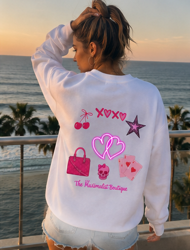 A woman stands on a balcony overlooking the ocean at sunset, wearing a white sweatshirt with a colorful back graphic featuring pink hearts, cherries, a handbag, cards, and the text 'The Maximalist Boutique'.