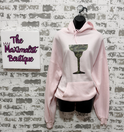 Pink hoodie with a graphic design on a textured wall with 'The Maximalist Boutique' sign.