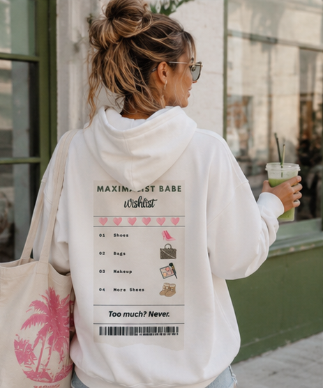 A woman viewed from behind wearing a white hoodie with a pink and black graphic print on the back, carrying a pink palm tree tote bag and holding an iced coffee in front of a storefront.