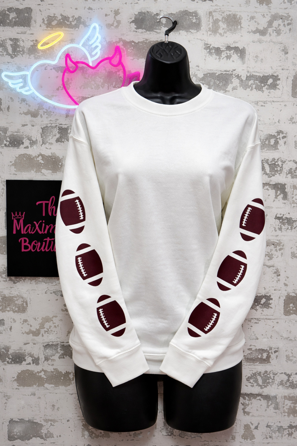 White sweatshirt with football designs on sleeves, displayed on a mannequin against a textured wall.